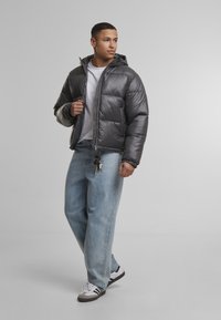 Pilkas pūkuotas švarkas su blizgiu paviršiumi ir kapišonu, derinamas su šviesiai mėlynomis džinsomis ir juodai baltais sportbačiais. Matomas raktų pakabukas.