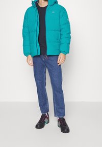 Teal puffjacka med huva, med en frontdragkedja och elastiska muddar. Matchad med mörka jeans och flerfärgade sneakers.