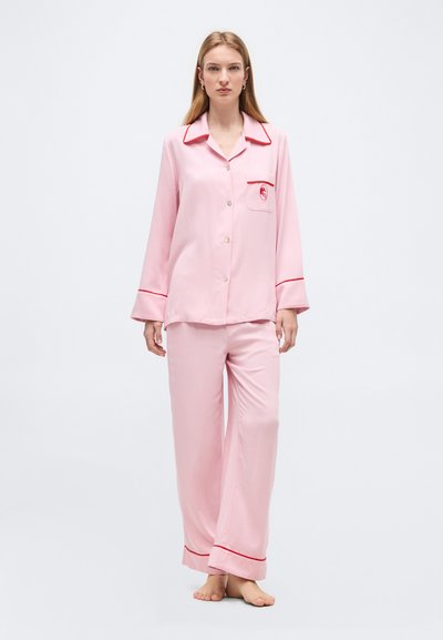 Γυναίκα στέκεται ξυπόλητη φορώντας ανοιχτό ροζ πυτζάμες με κόκκινη λεπτομέρεια στο γιακά, τις μανσέτες, την τσέπη και τα τελειώματα του παντελονιού, σε απλό λευκό φόντο.