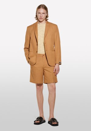 Brauner Leinenanzug mit einem taillierten Blazer und passenden Shorts, kombiniert mit einem hellgelben Strickshirt und schwarzen Sandalen.