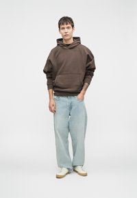 Hoodie castanho com um bolso frontal, corte solto. Combinado com jeans claros de perna larga e ténis em creme. Design simples e casual.