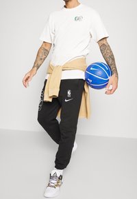 Basketbollspelare klädd i en vit t-shirt, svarta svettbyxor med NBA-logotyp och vita sneakers, håller i en blå basketboll. Ljus tröja knuten runt midjan.