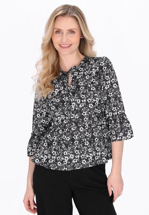 Smilende kvinde med langt blondt hår iført en sort og hvid blomsterbluse med 3/4-ærmer og sorte bukser på en hvid baggrund.