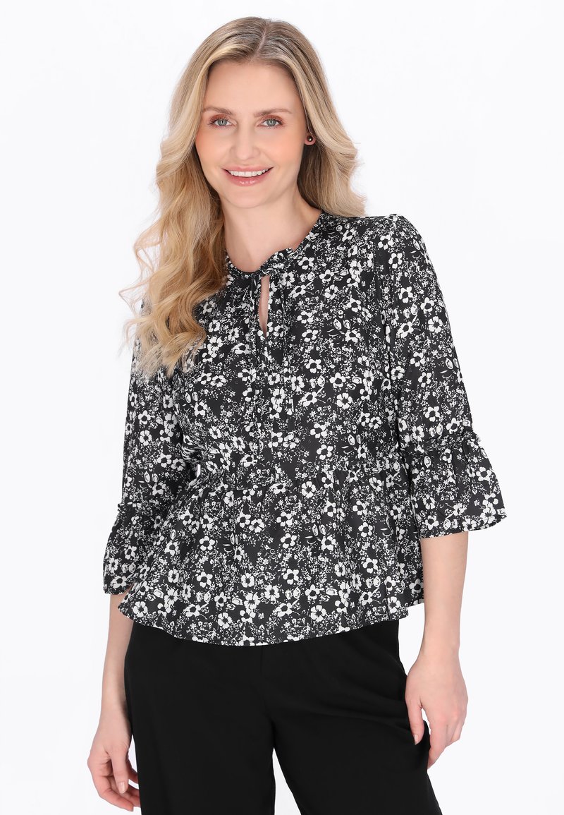 Femeie zâmbitoare cu păr lung și blond, purtând o bluză florală alb-negru cu mâneci 3/4 și pantaloni negri, pe un fundal alb.