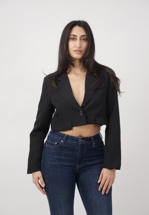 Frau mit langen dunklen Haaren, die ein schwarzes, kurzes Blazer und dunkelblaue Jeans trägt, steht vor einem einfarbigen hellgrauen Hintergrund.