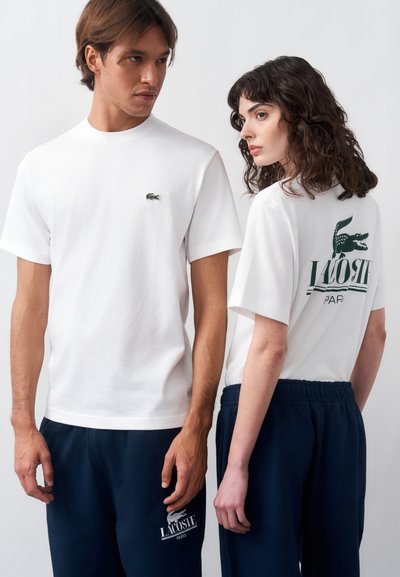 Мъж и жена, облечени в бели тениски Lacoste и тъмносини панталони, стоят един до друг на едноцветен фон.