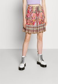 Jupe à motif floral en rose et beige avec taille à cordon, présentant un design évasé et des accents décoratifs au niveau de l'ourlet. Portée avec des bottes blanches.