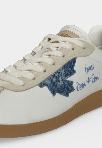Baskets blanches avec bordure en daim beige, lacets blancs, semelle gomme, patch en denim bleu en forme de feuille et texte bleu style manuscrit sur le côté.