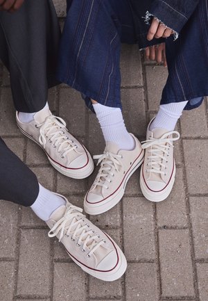 Trois personnes portant des baskets blanches avec des chaussettes blanches assises sur un sol pavé, montrant les jambes et les pieds.