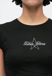 Zwart nauwsluitend t-shirt met een sterontwerp en de tekst "Guess Jeans" versierd met roze en witte strass-steentjes. Korte mouwen.