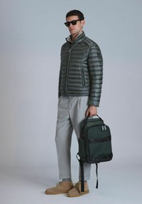 Veste rembourrée vert olive avec col haut, fermeture éclair et accents gris. Pantalons gris clair et bottes en daim marron. Sac à dos sombre avec des bretelles.