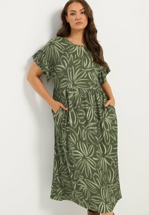 Femme portant une robe verte avec des motifs floraux clairs, des manches courtes volantées, et les mains dans les poches, debout contre un mur blanc.