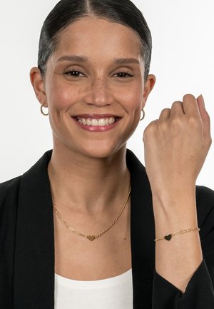Goldene Halskette mit Herzanhänger und passendem Armband, das aus verdrehten und verbundenen Kettendesigns besteht. Das Model trägt ein schwarzes Blazer und ein weißes Oberteil.