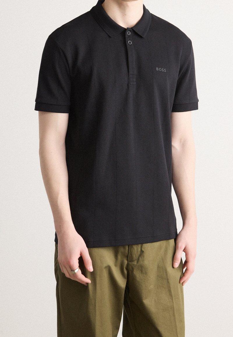 Homme portant un polo noir à manches courtes avec le logo "BOSS" sur la poitrine et un pantalon vert olive, debout devant un fond clair uni.