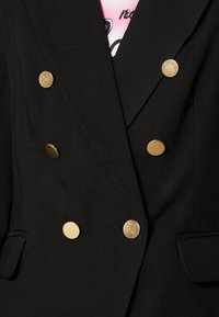 Musta bleiseri, jossa on kaksirivinen malli, kuusi pyöreää kultalangasta nappia ja kaksi etutaskua. Pehmeä kangas, jossa on kevyttä kiiltoa.