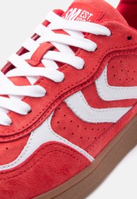 Κόκκινο σουέντ αθλητικό παπούτσι με λευκές δερμάτινες λεπτομέρειες, λευκά κορδόνια, διατρητές λεπτομέρειες και καφέ λαστιχένια σόλα, σε κοντινή όψη.