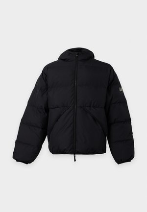 Homme debout sur fond blanc, portant une doudoune noire, un jean bleu délavé et des chaussures marron.