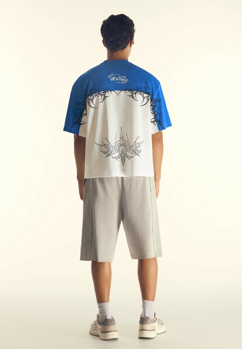 Man som står med ryggen mot kameran, klädd i en blå och vit kortärmad skjorta med svarta grafiska mönster, grå knälånga shorts, vita strumpor och sneakers.