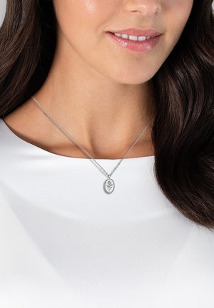 Collana con pendente ovale in argento con incisione floreale su una delicata catena. Indossata sopra una maglietta bianca, mette in risalto i dettagli del design.
