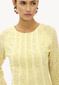 Top de encaje amarillo con recortes en forma de corazón, que presenta botones decorativos blancos a lo largo de las costuras verticales y un escote redondeado.