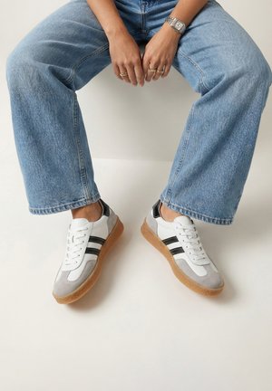 Osoba sedící, oblečená v světle modrých džínách a bílých teniskách s černými pruhy a gumovou podrážkou, ruce spočívající mezi nohama, na kterých jsou vidět prsteny a hodinky.