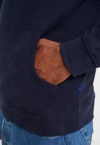 Felpa blu navy con tasca a marsupio, polsini e fondo a coste, e un piccolo etichetta blu del marchio di lato. Tessuto morbido.