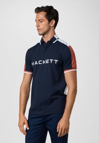 Marineblauwe polo met witte en oranje accenten, voorzien van een kraag, korte mouwen en het "HACKETT" logo op de voorkant. Glad stoftextuur.