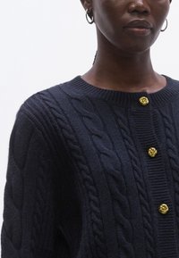 Καρντίγκαν σε σκούρο μπλε ναυτικό χρώμα με πλέξη καλωδίων και στρογγυλή λαιμόκοψη, διακοσμημένο με χρυσά σπειροειδή κουμπιά, υφασμένο σχέδιο και ριμπ λεπτομέρειες κατά μήκος των άκρων.