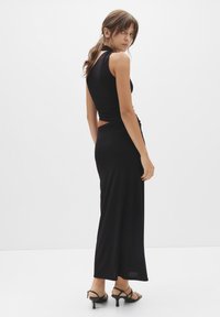 Schwarzes, figurbetontes Kleid mit einem Neckholder und offenem Rücken, das mit einem gerafften Seitendetail und einer langen, fließenden Silhouette versehen ist. Strapazierfähige schwarze High Heels.