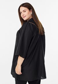 Žena s dlhými hnedými vlasmi, oblečená v voľnej čiernej textúrovanej blúzke s rukávmi po lakte, pozerajúca cez rameno, na plain bielom pozadí.