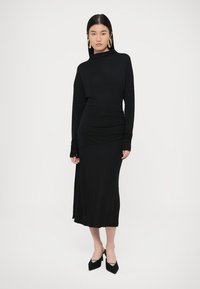 Fekete, hosszú ujjú ruha testhez simuló sziluettel, redőzött anyag részletekkel és magas nyakkal, fekete hegyes orrú magassarkúval párosítva.