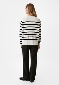 Maglione a righe beige e nero con orlo a coste, abbinato a pantaloni neri e scarpe basse. La vista posteriore mostra una vestibilità rilassata e maniche lunghe.