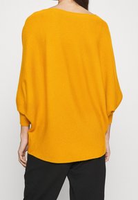 Femme portant un pull en tricot jaune moutarde à manches longues et ample, associée à un pantalon noir, vue de dos.