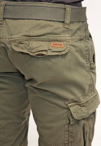 Pantalones cargo caqui con una cinturilla que incluye un cinturón texturizado. Tiene un bolsillo trasero con solapa y una etiqueta de marca de cuero.