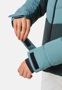 Dünne Jacke mit einem oberen Teil in Teal und einem unteren Abschnitt in Dunkelgrün. Verfügt über Klettverschlüsse an den Bündchen und ein schwarzes Innenfutter. Der Reißverschluss ist sichtbar.