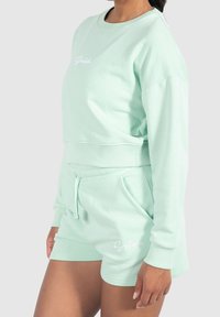 Felpa corta verde menta con polsini e orlo a coste, abbinata a pantaloni corti coordinati con cordoncino regolabile e dettaglio del logo.