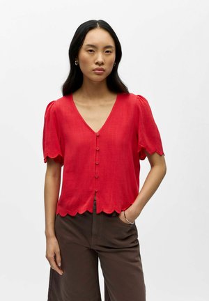 Femme portant un chemisier rouge à boutons avec des bords festonnés et des manches courtes, associé à un pantalon marron foncé, debout devant un fond uni.