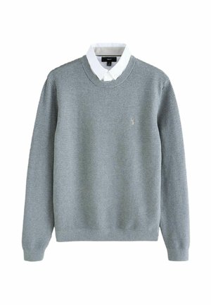 Pull en mailles texturé gris clair porté par-dessus une chemise blanche avec col, des manches longues, encolure ronde avec un petit logo brodé sur la poitrine.