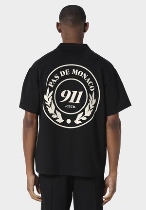 Mann, der von hinten zu sehen ist, trägt ein schwarzes Kurzarmshirt mit weißem, kreisförmigem "PAS DE MONACO 911 CLUB"-Logo und Lorbeerdesign auf dem Rücken.