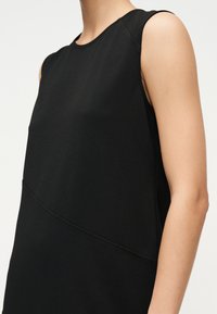 Μαύρο αμάνικο φόρεμα με στρογγυλή λαιμόκοψη και διαγώνια λεπτομέρεια ραφής, που φοριέται από ένα άτομο μπροστά σε απλό ανοιχτό φόντο.