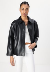 Μαύρο σακάκι από συνθετικό δέρμα με δομημένο σχέδιο, κλείσιμο με κουμπιά και φαρδύ γιακά. Φοριέται πάνω από ένα λευκό κοντό τοπ και τζιν.