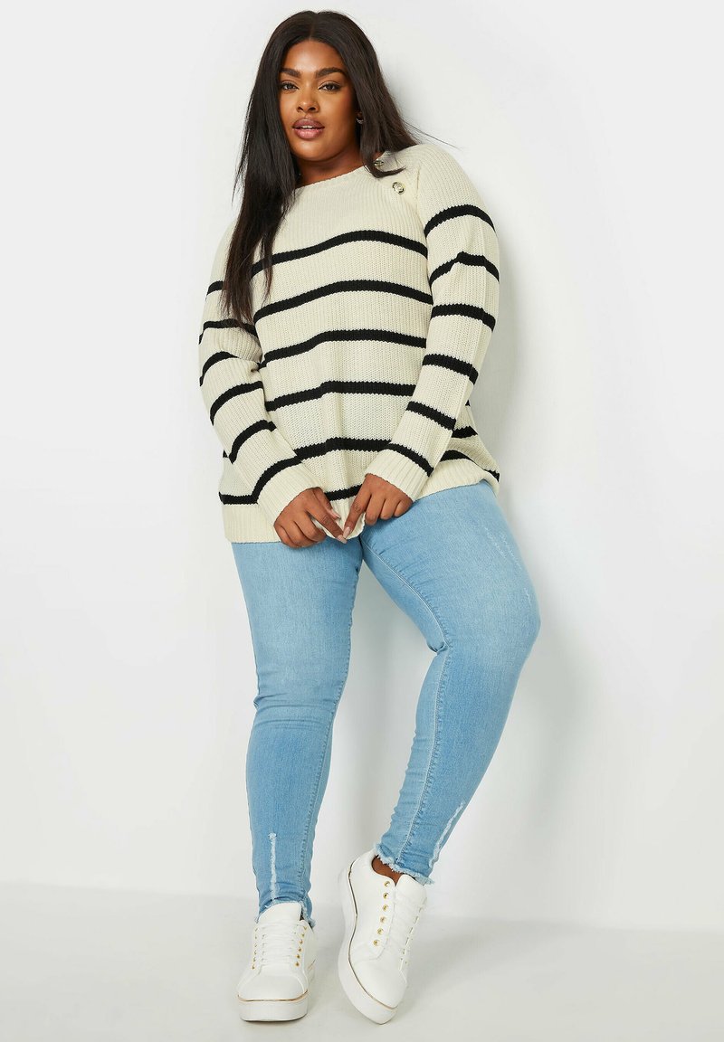 Krämfärgad ribbad tröja med svarta horisontella ränder, med rund halsringning. Kombinationen komplett med ljusblå smala jeans och vita sneakers.