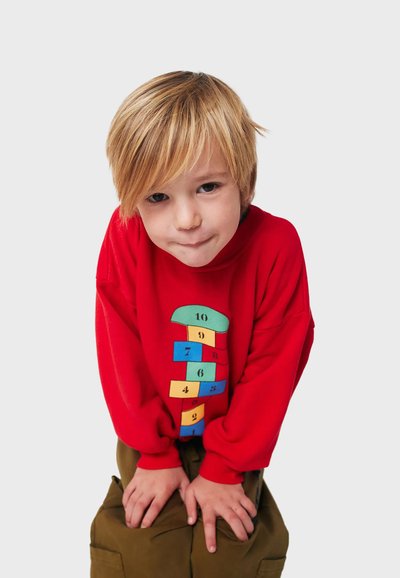 Felpa rossa con un design di campielli colorati che presenta numeri e forme geometriche. Maniche lunghe e vestibilità rilassata. Pantaloni marroni.