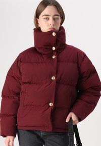 Burgunderfarbene Puffjacke mit hohem Kragen, goldenem Knopfverschluss, gestepptem Design und seitlichen Taschen. Getragen mit schwarzen Jeans und einem Tragegurt.