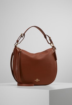 Brązowa torba typu hobo marki Coach ze skóry, z pozłacanymi elementami, regulowanym paskiem i małym logo z przodu, spoczywająca na białym piedestale na szarym tle.