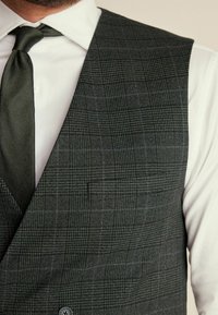 Šedá kostkovaná vesta s jemným vzorem a hladkým povrchem, kombinovaná s bílou košilí a tmavozelenou kravatou. Obsahuje detail kapsy.