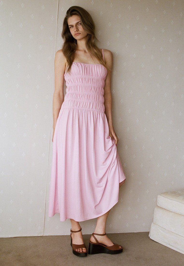 Jeune femme aux longs cheveux bruns portant une robe midi rose sans manches et des sandales compensées marron, debout à l'intérieur contre un mur à motifs.