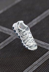 Zapatilla deportiva blanca con un patrón de camuflaje gris, que presenta una suela acolchada con acentos en azul verdoso y cordones texturizados, sobre un fondo de rejilla metálica.