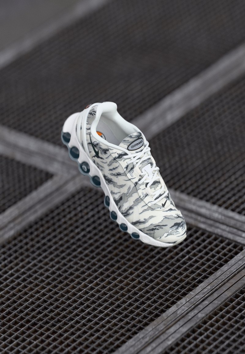 Zapatilla deportiva blanca con un patrón de camuflaje gris, que presenta una suela acolchada con acentos en azul verdoso y cordones texturizados, sobre un fondo de rejilla metálica.