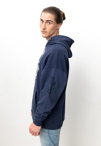 Marineblaue Kapuzenjacke mit Kängurutasche, Zugband-Haube und Grafik auf der Vorderseite. Mit aufgedrucktem Logo auf dem Ärmel. Aus weichem Baumwollmaterial.